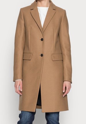 Mujer con un abrigo de lana color camel marrón hasta la rodilla, con dos botones, bolsillos con solapa y cuello de muesca, sobre una camiseta blanca y vaqueros.