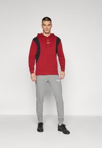 Felpa rossa con accenti neri e un logo, abbinata a pantaloni felpati grigi con un logo bianco, completata da scarpe da ginnastica nere.