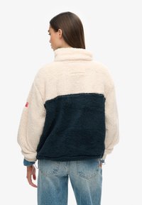 Kurtka z polaru z przytulnym kremowym korpusem i ciemnoniebieskim panelem. Wysoki kołnierz, oversize'owe rękawy i regulowany dół. Prosty czerwony akcent w formie logo.