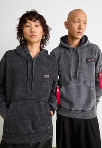 Zwei Personen tragen dunkelgraue Kapuzenpullover mit einer leicht abgenutzten Textur. Die Kapuzenpullover verfügen über eine Vordertasche und Markenaufnäher sowie rot gefärbte Reißverschlussakzente.