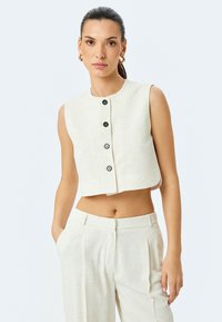 Gekürztes beiges Leinen-Hemd ohne Ärmel mit rundem Halsausschnitt und fünf schwarzen Knöpfen auf der Vorderseite. Schlichte Textur mit minimalistischem Design.
