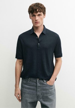 Giovane uomo con polo blu scuro a maniche corte infilata in jeans di denim grigi, in piedi con una mano in tasca.