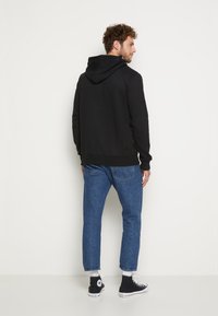 Schwarzer Kapuzenpulli mit weicher Textur, lässigem Schnitt und gerippten Bündchen, kombiniert mit hellblauen, schmal geschnittenen Jeans und schwarzen High-Top-Sneakern.