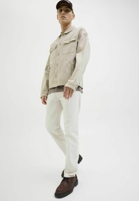 Giacca di denim beige chiaro con due tasche sul petto, indossata sopra una camicia grigia chiara e pantaloni bianchi. Scarpe stringate marroni con suola texture.