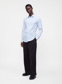 Uomo in piedi che indossa una camicia azzurro chiaro con bottoni, pantaloni neri a gamba larga e scarpe nere su uno sfondo bianco semplice.
