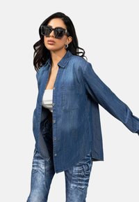 Hellblaue Denim-Hemd mit Knopfleiste, lange Ärmel, eine Brusttasche, weiche Textur, kombiniert mit dunkelblauen Denim-Hosen. Schwarze quadratische Sonnenbrille getragen.
