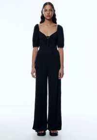 Schwarzer Jumpsuit mit quadratischem Ausschnitt, Puffärmeln und Bindeband vorne. Hergestellt aus strukturiertem Stoff, mit weiten Hosenbeinen, kombiniert mit Plateausandalen.