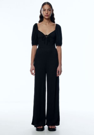Zwarte jumpsuit met een vierkante halslijn, pofmouwen en strikdetail aan de voorkant. Gemaakt van textuurstof, met een wijde broekspijp, gedragen met plateau-sandalen.