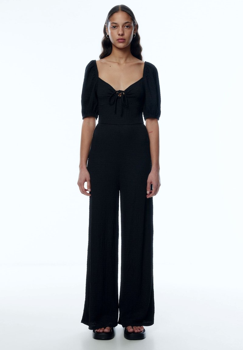 Schwarzer Jumpsuit mit quadratischem Ausschnitt, Puffärmeln und Bindeband vorne. Hergestellt aus strukturiertem Stoff, mit weiten Hosenbeinen, kombiniert mit Plateausandalen.