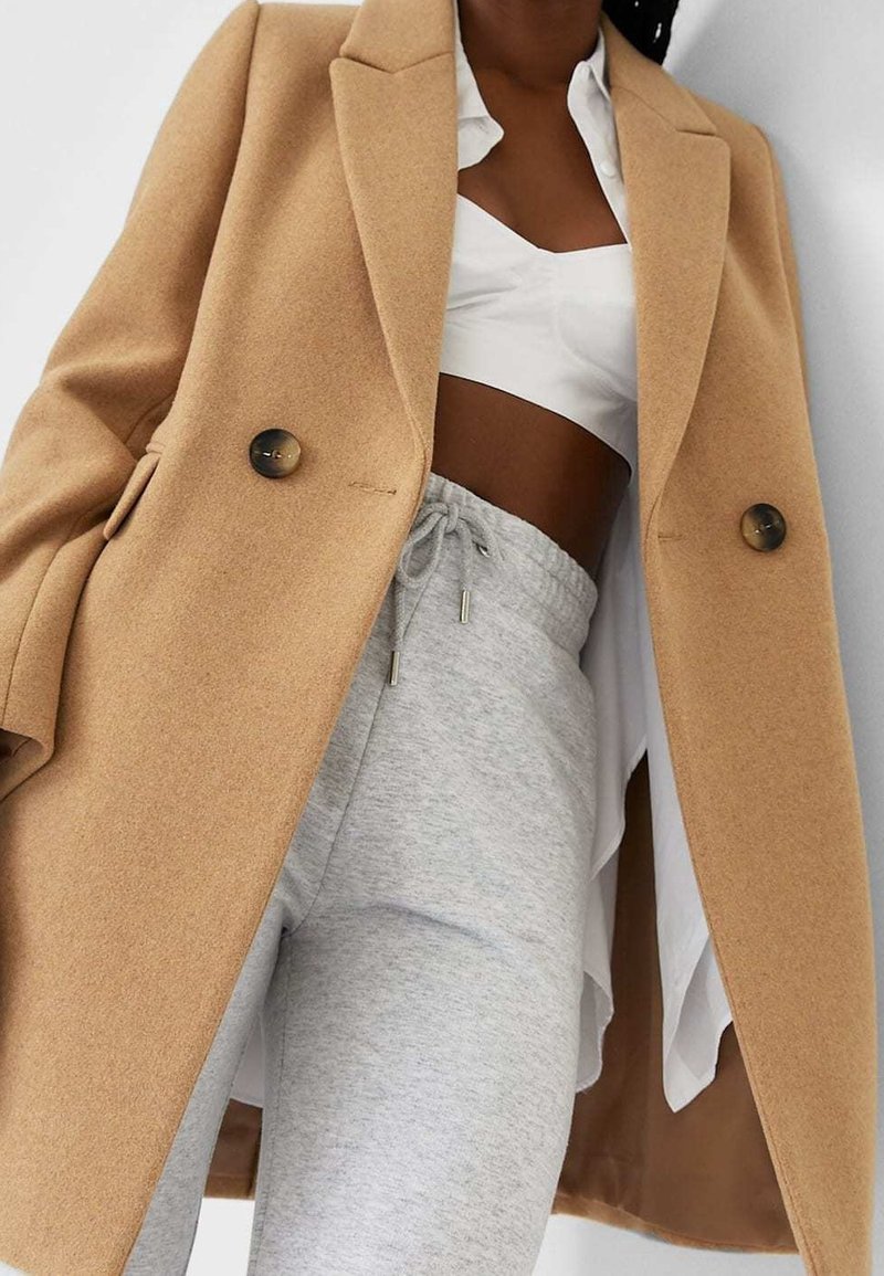 Femme portant un manteau croisé couleur camel sur un top court blanc et un pantalon de jogging gris clair avec cordon, sur un fond uni.
