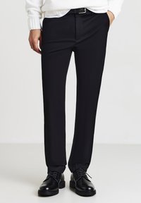 Pantaloni neri eleganti con una vestibilità slim, tessuto liscio e una leggera lucentezza. Abbinati a scarpe in pelle nere e un maglione bianco a maglia.