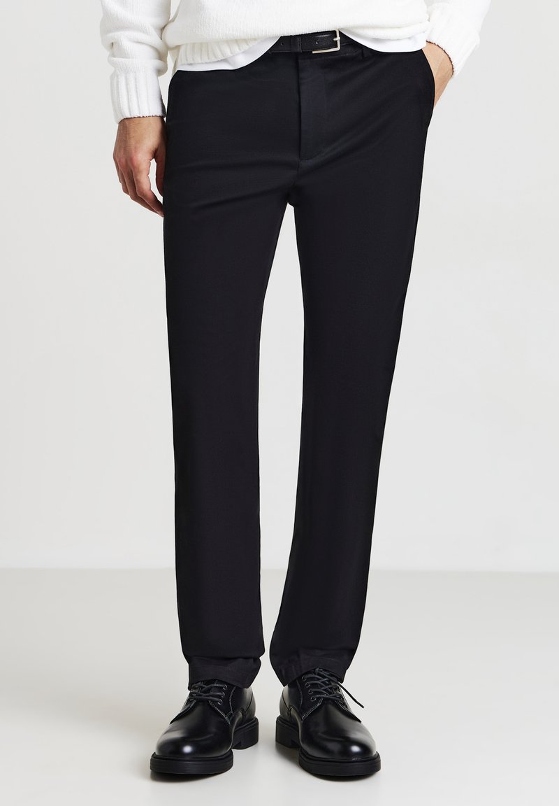 Pantaloni neri eleganti con una vestibilità slim, tessuto liscio e una leggera lucentezza. Abbinati a scarpe in pelle nere e un maglione bianco a maglia.