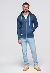 Granatowa bluza z kapturem na zamek, z białymi sznurkami, w połączeniu z jasnoniebieskimi dżinsami i żółtymi trampkami, stojąca na neutralnym tle.