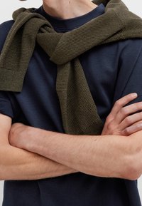 Laivastonsininen lyhythihainen paita, jonka päällä on teksturoitu tummanvihreä neulepusero hartioilla, kädet ristissä edessä.