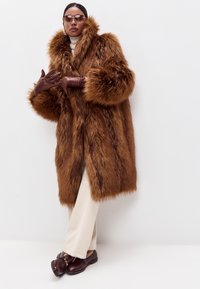 Brun pelsjakke med en stor krave og brede ærmer, parret med beige bukser og mørkebrune sko. Modellen bærer brune handsker og solbriller.