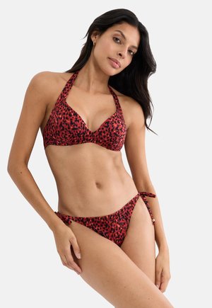 Femme portant un haut de bikini licou à imprimé léopard rouge et noir et un bas de bikini assorti à nouer sur les côtés, debout avec la main sur la hanche devant un fond blanc.