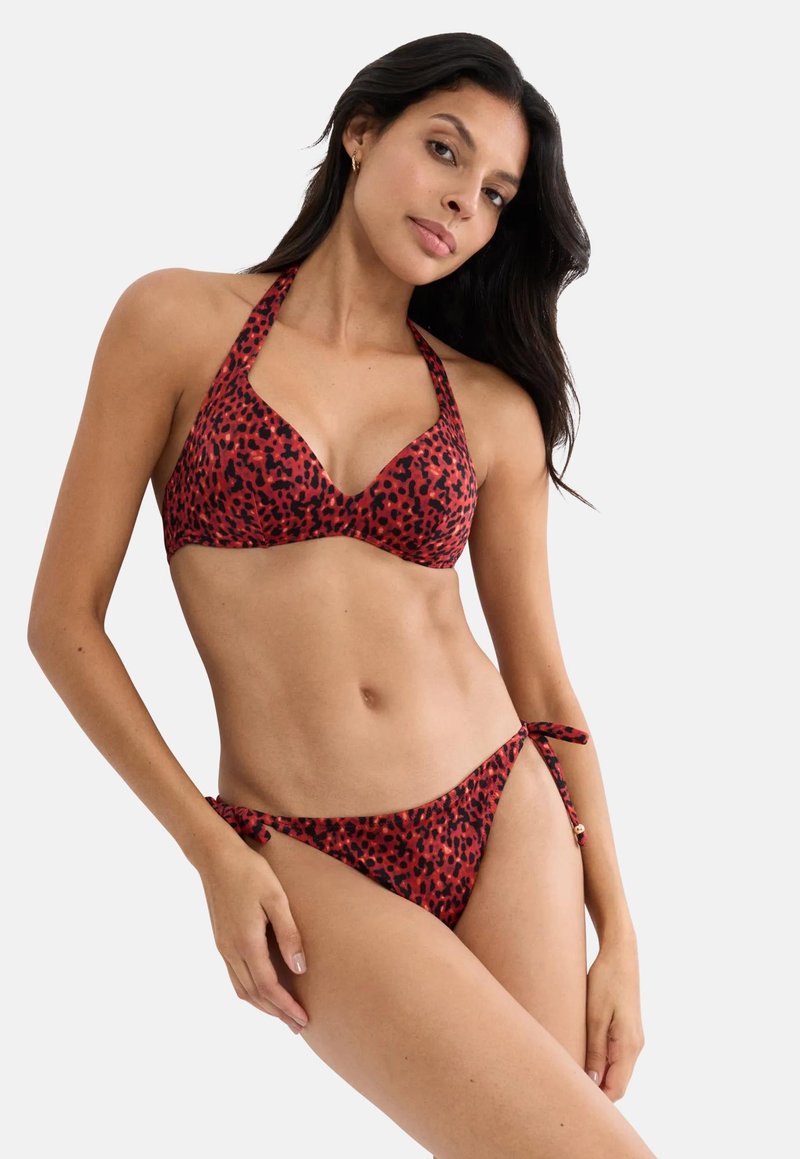 Femme portant un haut de bikini licou à imprimé léopard rouge et noir et un bas de bikini assorti à nouer sur les côtés, debout avec la main sur la hanche devant un fond blanc.