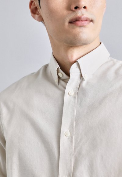 Camisa color beige claro de botones con un cuello clásico, hecha de una tela suave. Presenta costuras visibles y botones claros.
