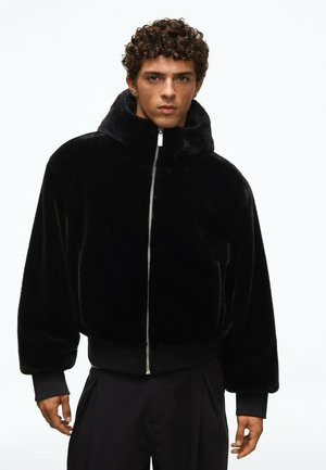 Jeune homme aux cheveux bouclés portant une veste en peluche noire zippée avec un col haut et des poignets élastiques, debout devant un fond uni.