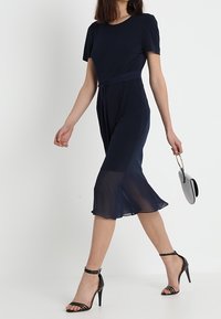 Vestido azul marino con mangas cortas y un dobladillo acampanado. Parte inferior de tela transparente con una cintura ajustada. Combinado con tacones negros de tiras y un bolso pequeño.
