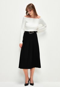 Svart plisserad midi kjol med en definierad midja, kombinerad med en vit ribbad tröja som är off-the-shoulder. Komplett med svarta klackskor.