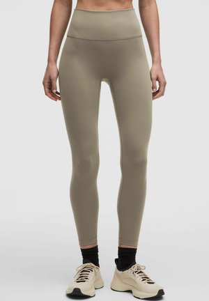 Donna che indossa leggings beige a vita alta, calzini neri e scarpe sportive beige, in piedi su sfondo chiaro.