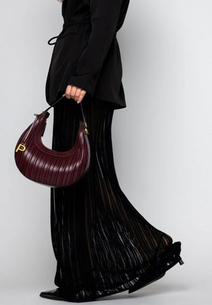 Burgunderfarbige Lederhandtasche mit plissiertem Design und goldenen Beschlägen, gehalten neben einem langen schwarzen, transparenten, plissierten Rock und spitzen schwarzen Schuhen.