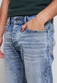 Homme portant un jean bleu clair délavé et une chemise vert foncé, main droite dans la poche avant.