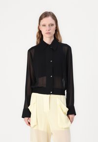 Camicia nera trasparente con colletto, maniche lunghe e orlo elastico; abbinata a pantaloni palazzo giallo chiaro a vita alta con ampie tasche.