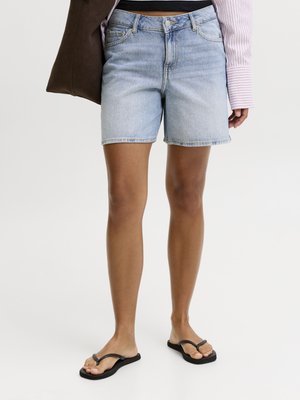 Persona que lleva pantalones cortos de mezclilla azul claro, chanclas negras, sostiene una bolsa marrón oscuro y lleva una camisa de manga larga rosa con rayas.