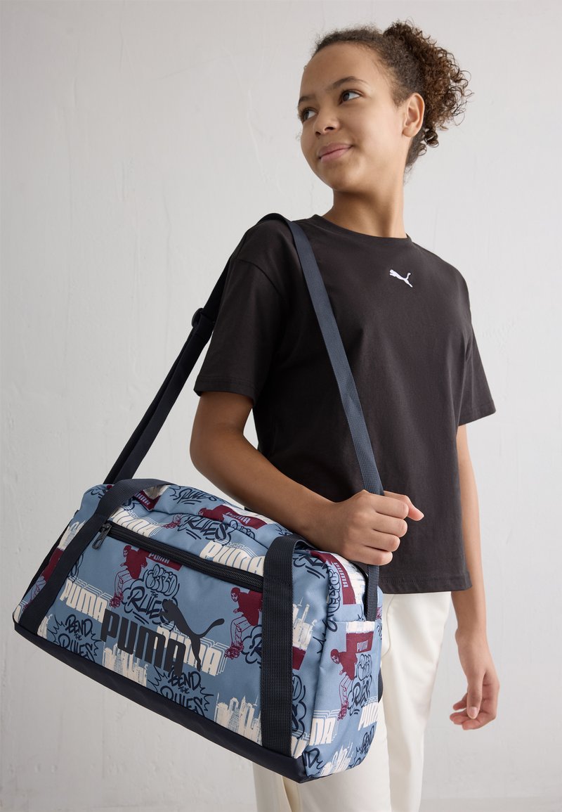Kék mintázattal rendelkező sporttáska, piros és fehér grafikával. Tartós anyagból készült, sötét pántokkal és cipzáras zsebbel rendelkezik.