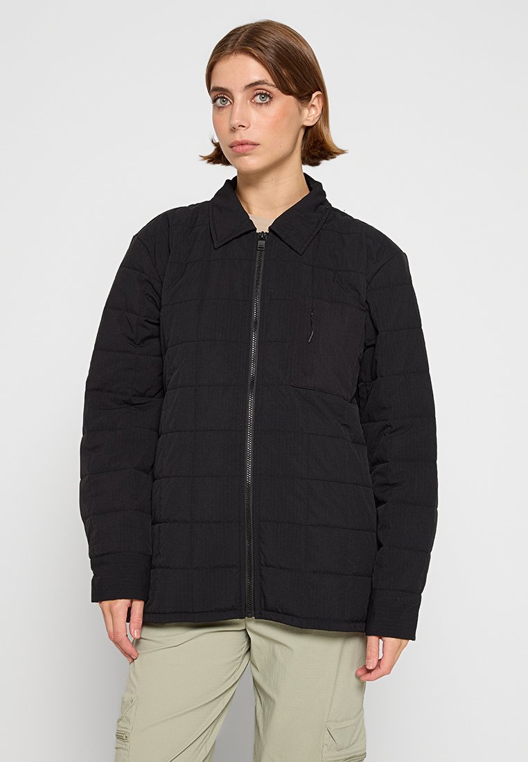 Kvinna med kort brunt hår klädd i en svart kviltad zip-up jacka med en bröstficka och ljusbeiga cargobyxor, stående mot en enkel bakgrund.