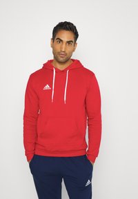 Roter Kapuzenpullover mit weißen Kordeln, Fronttasche und weißem Adidas-Logo auf der linken Brust. Kombiniert mit dunkelblauen Hosen.