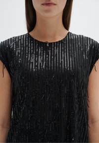 Černé šaty s krátkými rukávy, které mají vertikální pruhy z flitrů a texturované akcenty; látka má hladký lesk.