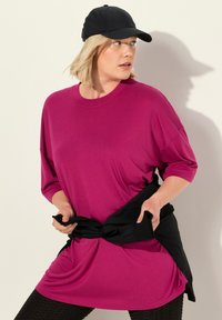 Overmaatse magenta T-shirt met driekwartmouwen, gecombineerd met een zwarte knoopshirt om de taille en textuur zwarte leggings.