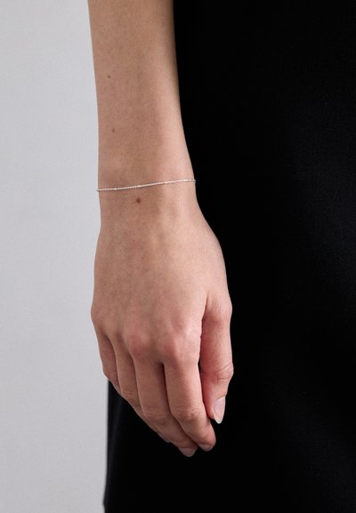 Bracelet en chaîne argentée porté sur un poignet, présentant un design délicat avec de petits maillons ronds, affiché sur un fond sombre.