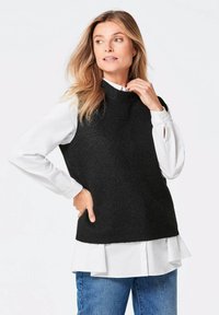 Gestrickter schwarzer ärmelloser Pullover, der über einem weißen Hemd mit Schlitz an den Manschetten getragen wird, kombiniert mit blauen Jeans. Glatte Textur, hochgeschnittenes Design.