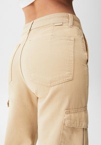 Pantalon cargo beige avec une taille ajustée, une grande poche latérale et une poche arrière au design carré. Tissu en coton doux avec une texture lisse.