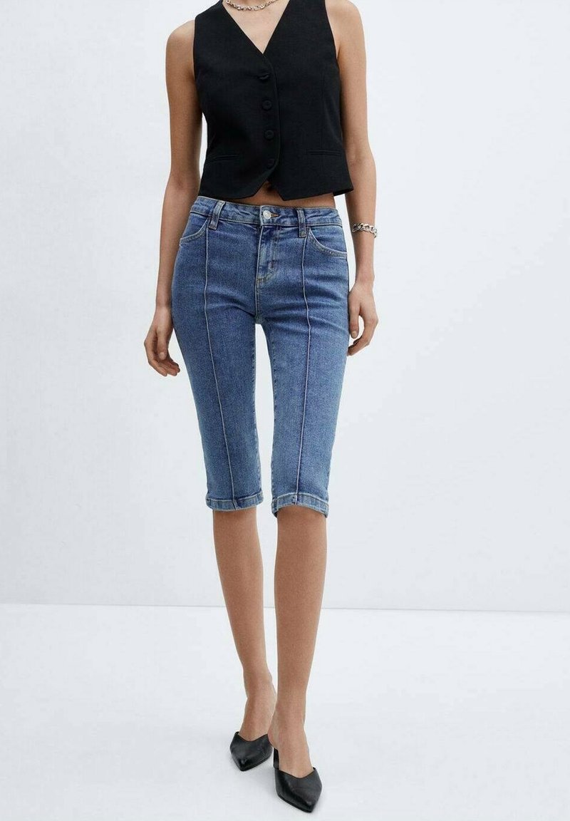 Jasnoniebieskie dżinsowe spodnie capri z wysokim stanem, pięcioma kieszeniami i kontrastowym szwem. W zestawieniu z czarną bluzką bez rękawów.