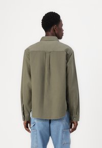 Olīvu zaļš garām piedurknēm krekls ar aizmugurējo šuvju un pogojamām aprocēm. Gluda auduma ar brīvu piegriezumu, ar sānu šķēlumiem zemē.