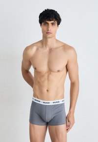 Boxers en gris avec une ceinture blanche arborant le logo "HUGO". Le modèle a une silhouette musclée et se tient devant un fond uni.