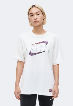 Młoda osoba z krótkimi blond włosami nosi białą, oversize'ową koszulkę Nike United oraz czarne legginsy z logo Nike na udzie.