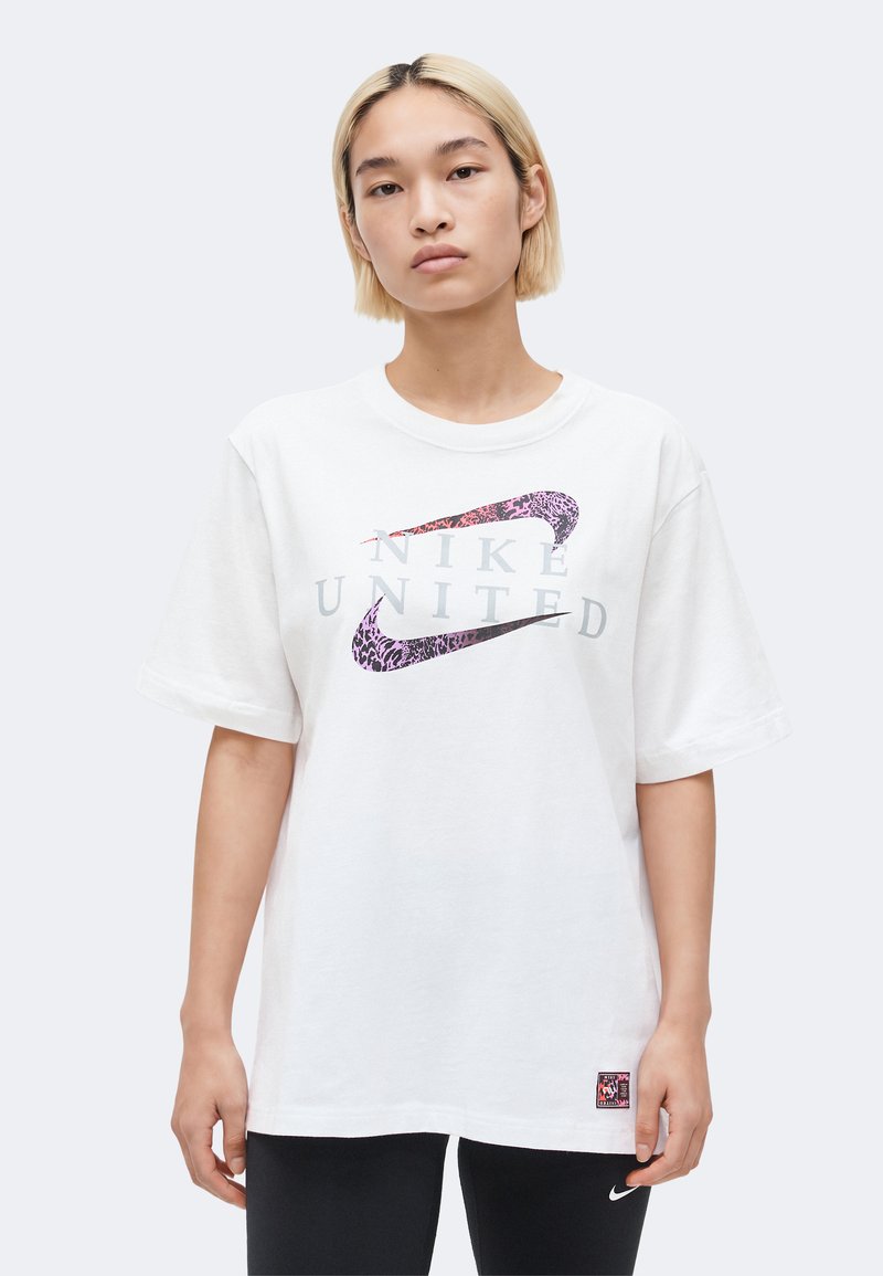 Νεαρό άτομο με κοντά ξανθά μαλλιά φοράει λευκό φαρδύ μπλουζάκι Nike United και μαύρο κολάν με λογότυπο Nike στον μηρό.