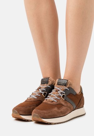 Baskets en daim marron et en shearling avec des lacets blancs à motifs, portées sur des jambes nues, avec une semelle blanche et des accents noirs.