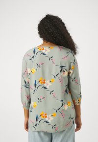Donna con lunghi capelli ricci che indossa una blusa floreale verde chiaro a tre quarti e jeans azzurri, vista da dietro.