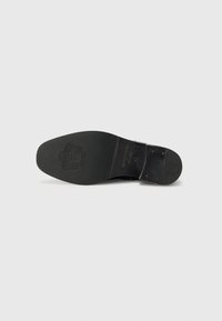 Semelle de chaussure en cuir noir avec une surface lisse, un bout arrondi et un logo en relief. Dotée d'un attache en métal au talon et d'une adhérence texturée.