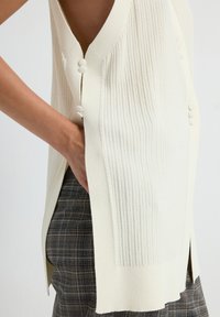 Chaleco de punto color marfil con textura acanalada, aberturas laterales y botones blancos redondos. Combinado con pantalones oscuros a cuadros.
