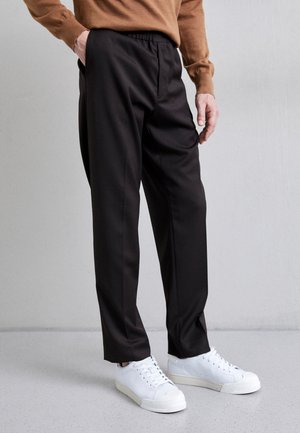 Personne portant un pantalon noir à jambes droites avec taille élastique et des baskets blanches, une main dans la poche, debout sur un sol gris clair.