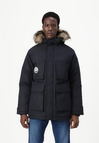 Parka nera imbottita con cappuccio foderato in pelliccia, molteplici tasche frontali e chiusura a scatto. Texture liscia con un materiale resistente e impermeabile.