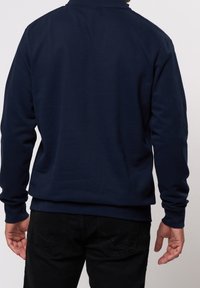 Mörkblå sweatshirt med hög krage, ribbade muddar och slät textur. Bärs tillsammans med svarta byxor, vilket visar en avslappnad stil.